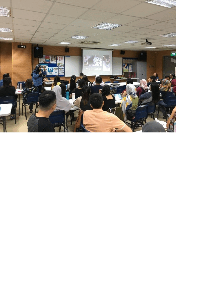 A group of people sit at tables in a classroom facing a projected presentation screen at the front.